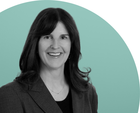 Professional black-and-white headshot of a woman with shoulder-length dark hair, wearing a dark blazer over a black top, smiling at the camera against a teal gradient background.