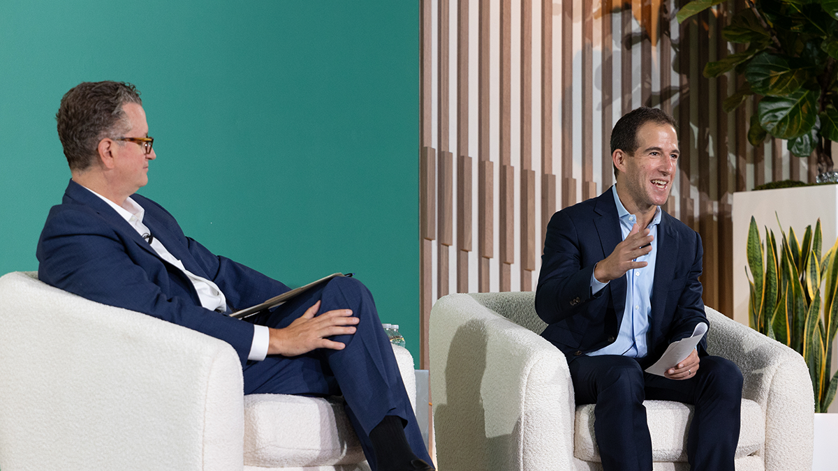 Apollo 2025 CEO Conference Panel Dialogue Two executives in navy suits speak during the 2025 Apollo CEO Conference, seated in white armchairs on stage, with modern wood paneling and greenery in the background.