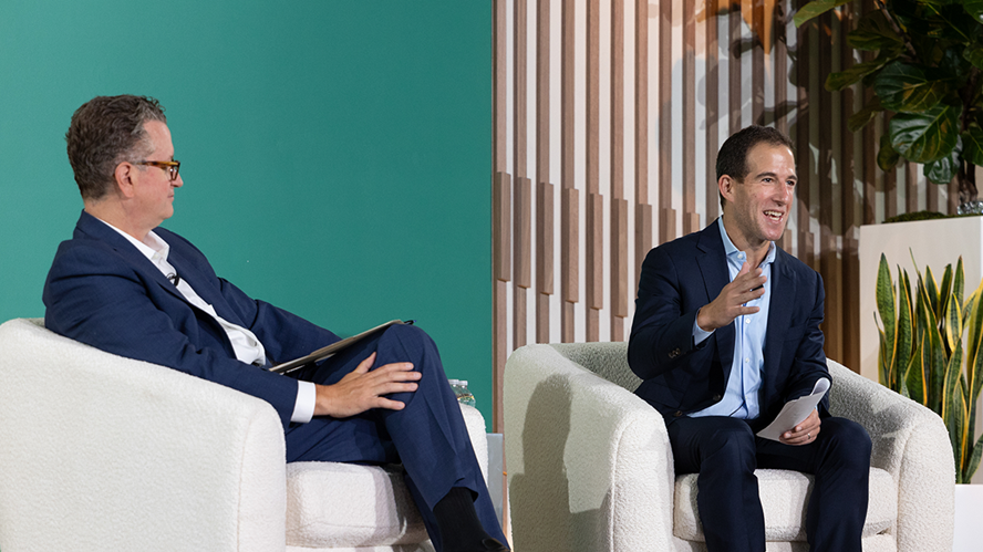 Two executives in navy suits speak during the 2025 Apollo CEO Conference, seated in white armchairs on stage, with modern wood paneling and greenery in the background.