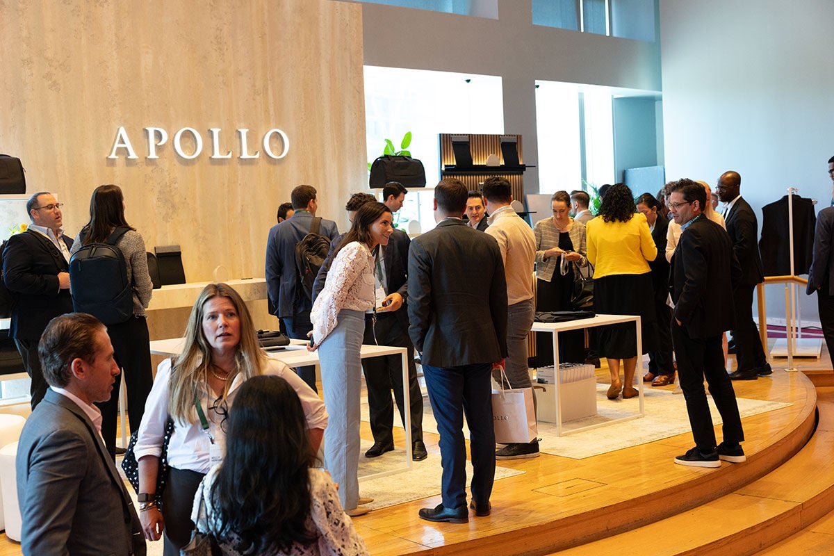Attendees networking during a break at an Apollo event, gathered in a bright open space with the "APOLLO" logo on a wood-paneled wall in the background.