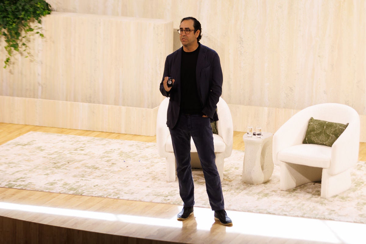 John Zito speaking on stage at an Apollo event, standing in front of the green 'APOLLO' logo on a stone backdrop, with white lounge chairs and a potted plant behind him."