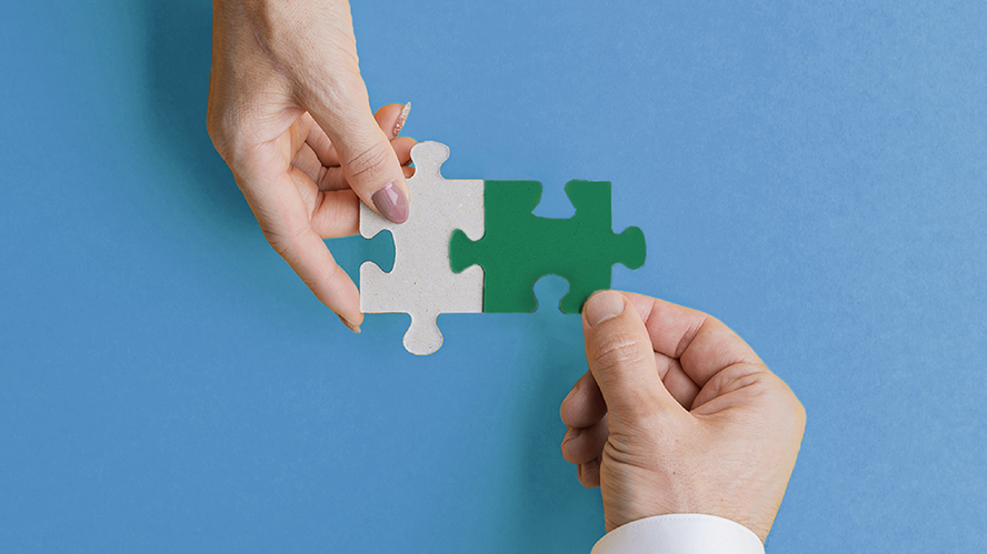 Two hands connect white and green puzzle pieces against a blue background, symbolizing collaboration, partnership, alignment, and shared problem-solving through complementary solutions and teamwork.