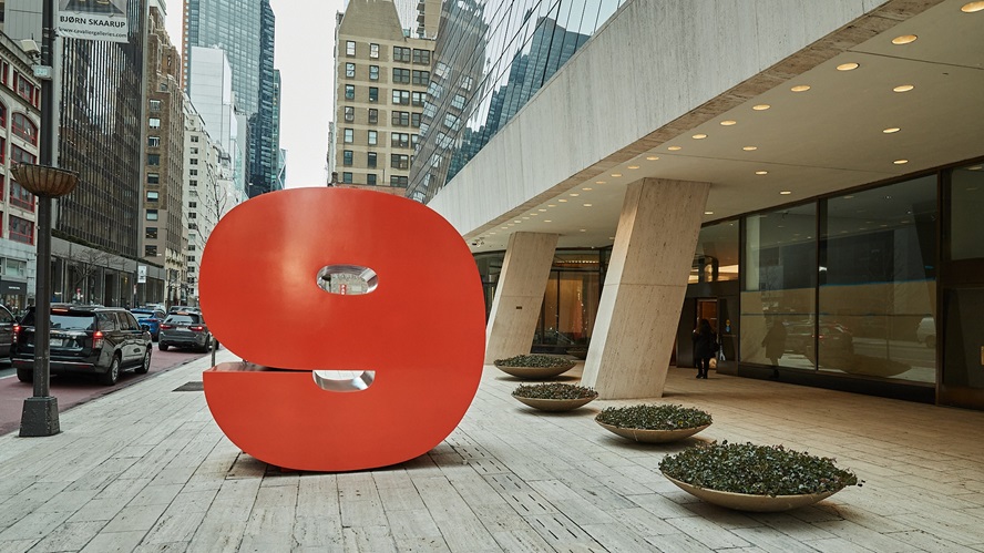 Large red number nine sculpture displayed on a pedestrian plaza outside a modern office building in an urban business district, with high-rise buildings, parked vehicles, and angled architectural columns framing the streetscape.