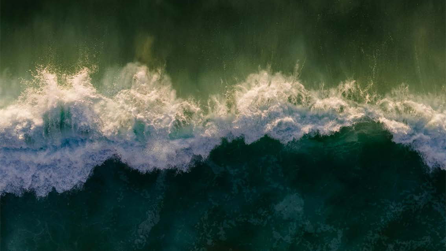 Aerial view of ocean waves breaking across deep green water, with white sea foam creating textured patterns along the surface. The image conveys the scale, movement, and natural energy of a coastal marine environment.
