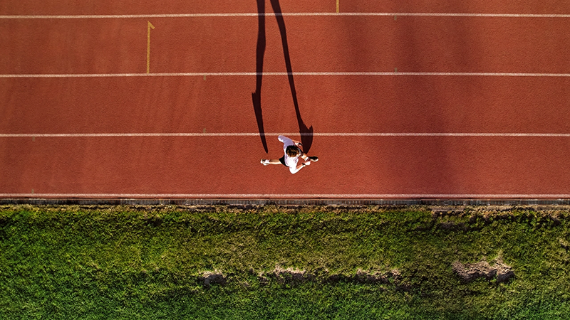 The Financing Gap in Sports: Unlocking a $2.5 Trillion Opportunity Overhead view of a runner in white athletic gear sprinting on a red track near a grassy field, casting a long shadow in the early morning light.