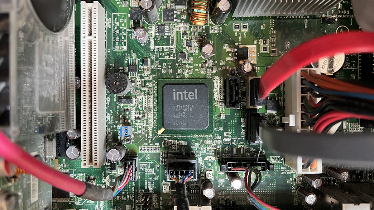 Close-up view inside a desktop computer showing a green motherboard with an Intel chipset, expansion slots, capacitors, heat sink, power cables, and data connections mounted on the circuit board.