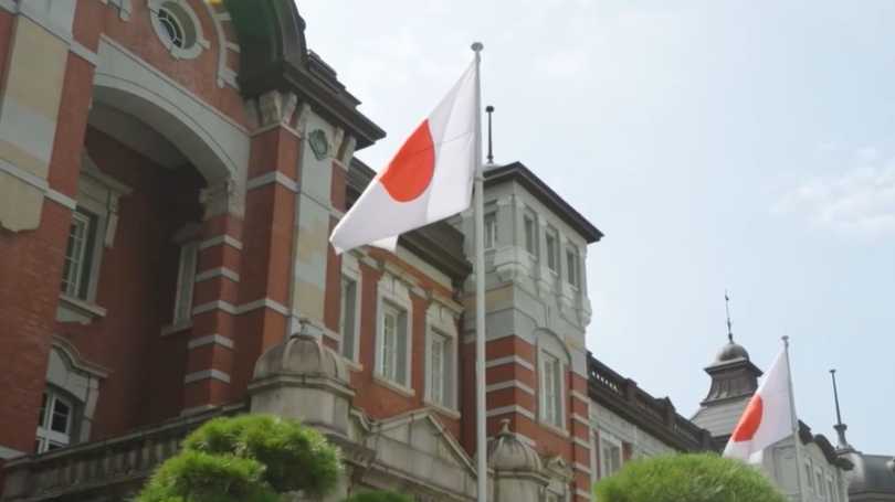 Japanese flags displayed outside a historic red-brick building with modern office towers in the background, representing Tokyo’s financial district and global capital markets activity in an urban setting.