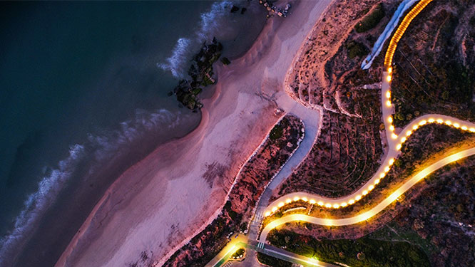 Aerial night view of a winding coastal road illuminated by golden streetlights, running alongside a sandy beach and dark ocean waters.