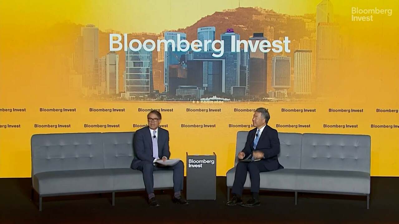 Wide view of a Bloomberg Invest conference stage with two speakers seated on gray sofas against a backdrop featuring the Hong Kong skyline and Bloomberg Invest branding during a live panel discussion on financial markets.