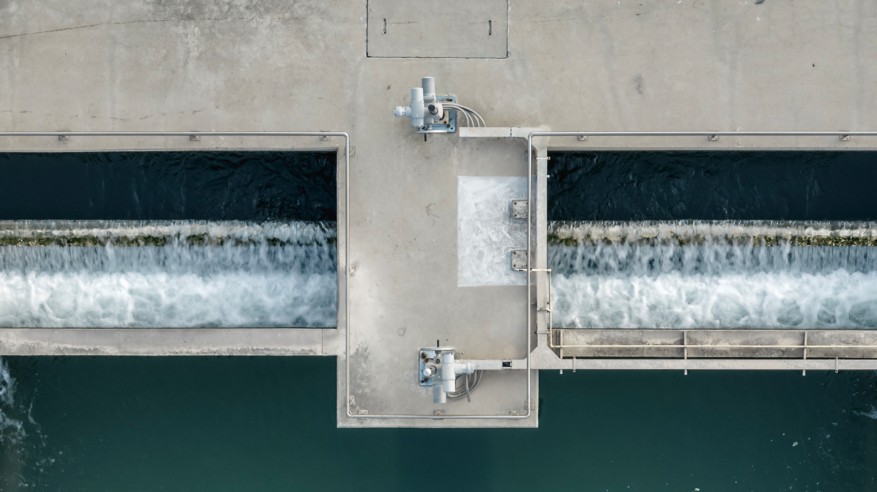 Aerial view of a dam structure with two parallel water outlets releasing white, foaming water over concrete weirs into a teal-green reservoir below.