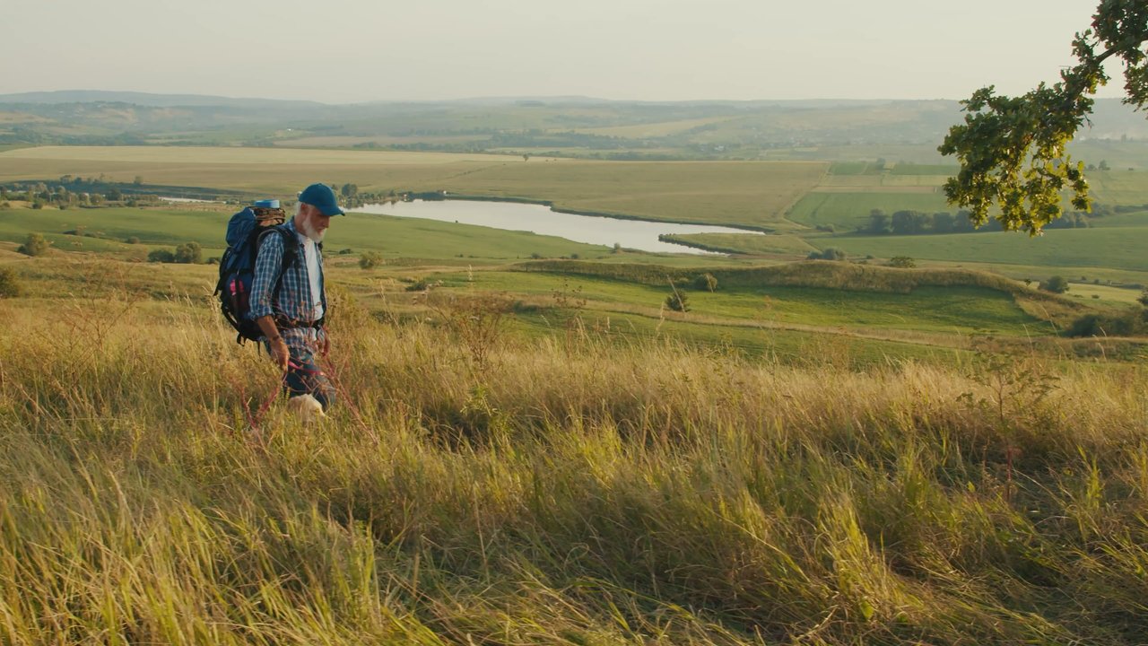 Older hiker with a backpack walks through tall grass on a hillside overlooking farmland and a winding river, illustrating an active retirement in a rural landscape.