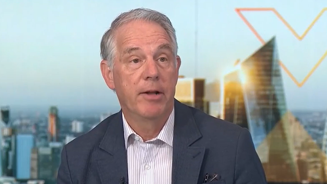 Jim Zelter speaks during a Bloomberg television interview, wearing a navy blazer and light striped shirt, with a blurred city skyline and studio graphics in the background as he discusses financial markets and investment topics.