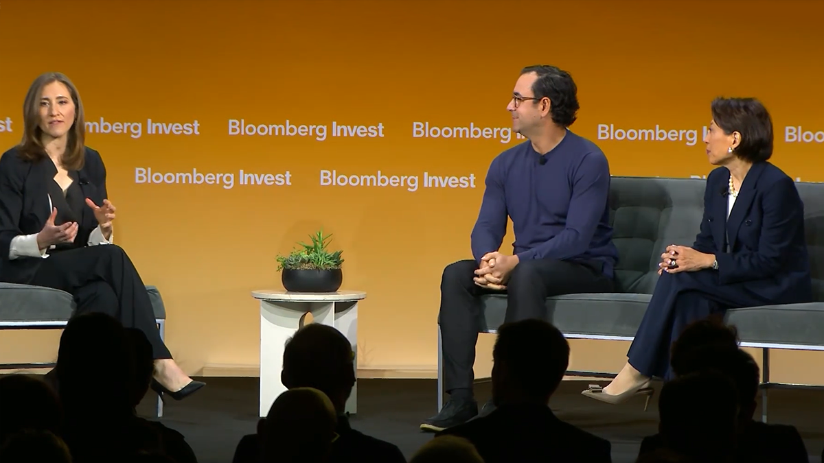 Panel discussion at Bloomberg Invest featuring three speakers seated on stage in front of a orange backdrop with the Bloomberg logo discussing private credit and retail access to private markets, with audience silhouettes in the foreground.