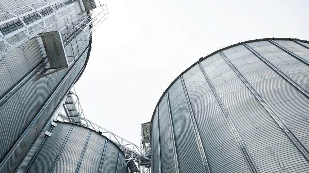 Upward view of large industrial storage tanks and metal infrastructure, representing hard assets infrastructure supporting AI deployment, including data centers factories, fiber, railroads. 