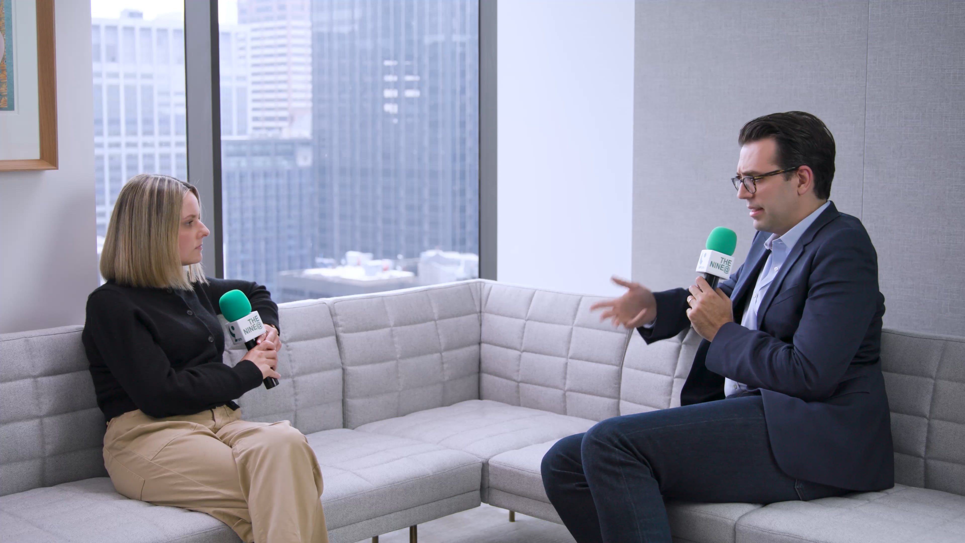 Paul Sipio sits on a modern office sofa during a 9@9 interview, holding a green microphone, speaking with a host against a city skyline backdrop. The discussion centers on private credit, capital markets, and asset management insights.