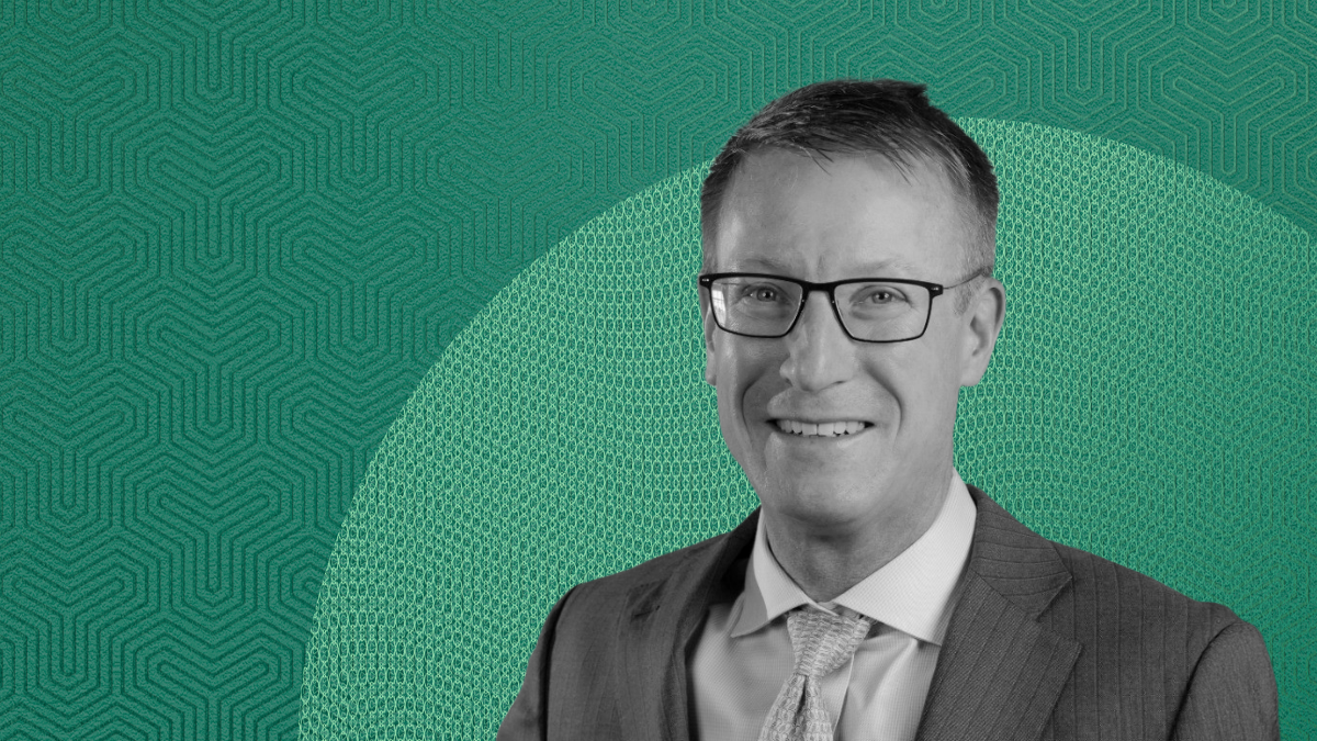 Black-and-white portrait of Mike Downing in a striped suit and glasses, smiling in front of a teal patterned background, discussing retirement income strategies.