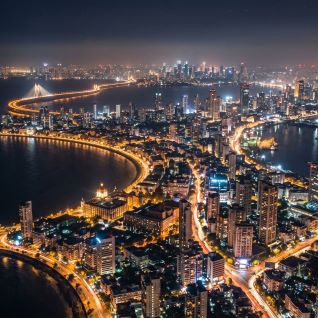 Aerial nighttime view of Mumbai’s coastline with illuminated highways curving along the waterfront, dense high-rise buildings, and the city skyline extending into the distance, illustrating a major global financial center.