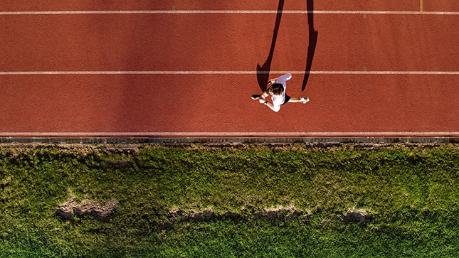 Overhead view of a person running on a red athletics track beside green grass, casting a long shadow in early morning or late afternoon light.