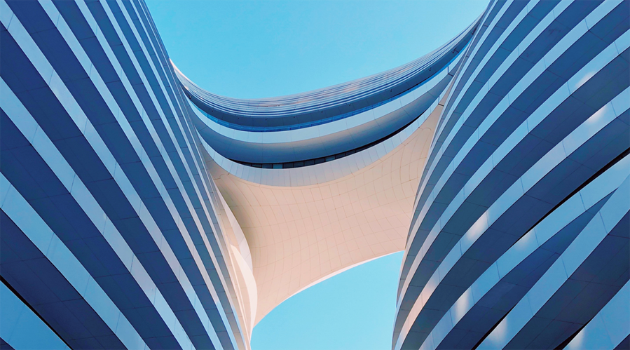 Upward view between two modern office towers connected by a curved architectural structure against a clear blue sky, illustrating contemporary commercial real assets and large-scale urban development.